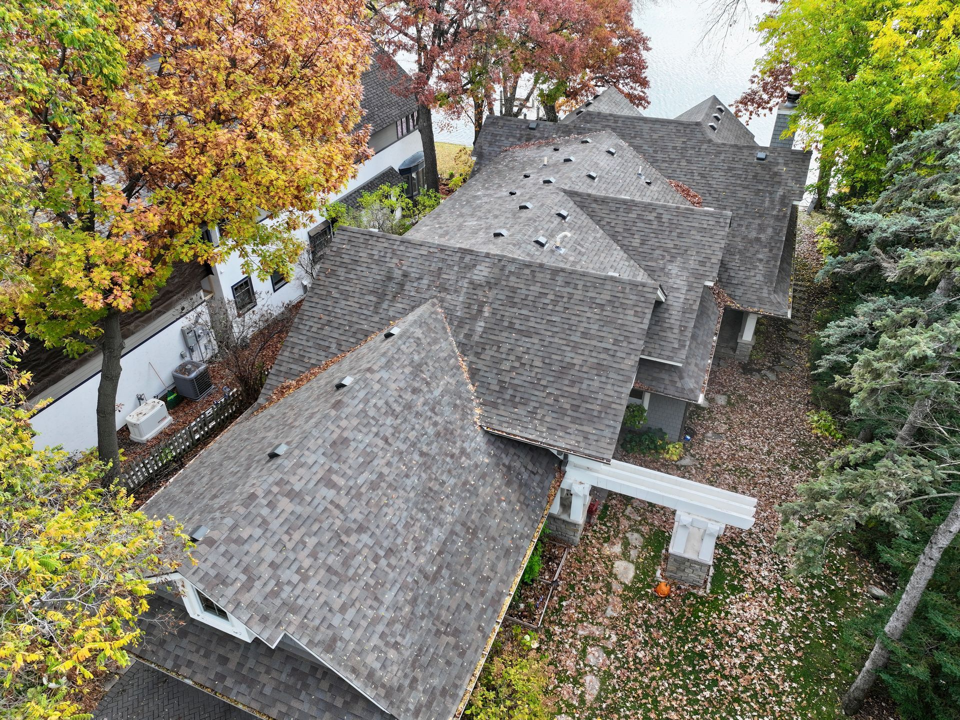 Overhead view of a house with multiple gray roofs, surrounded by trees with colorful fall foliage.