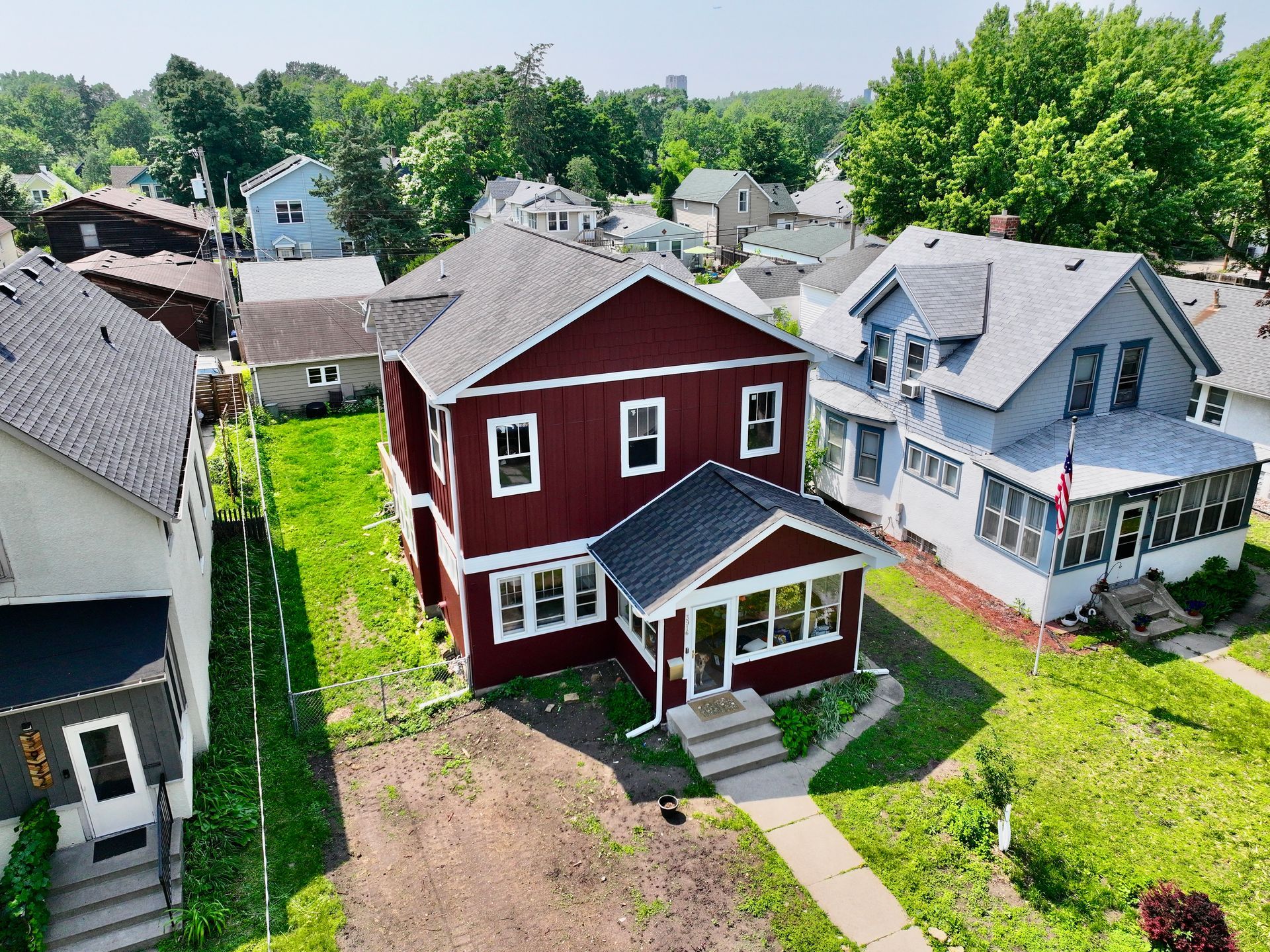 Red two-story house with white trim; houses and green trees in the background. Sunny day.