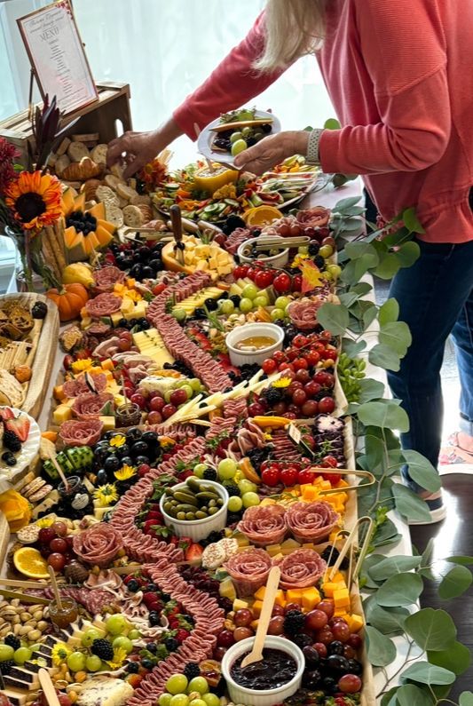 A large grazing table overflowing with food, including cheese, fruit, and charcuterie. A person reaches to arrange items.