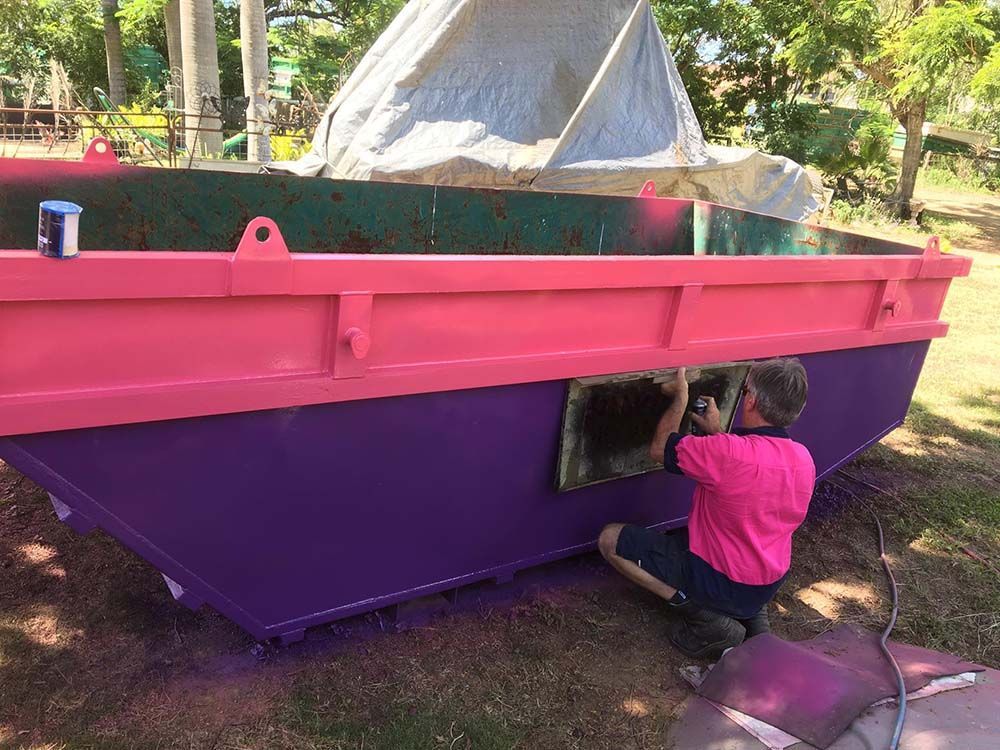 Man In A Pink Shirt Painting A Name On Pink And Purple Dumpster — Kanga Bins Qld Pty Ltd In Mulara, QLD