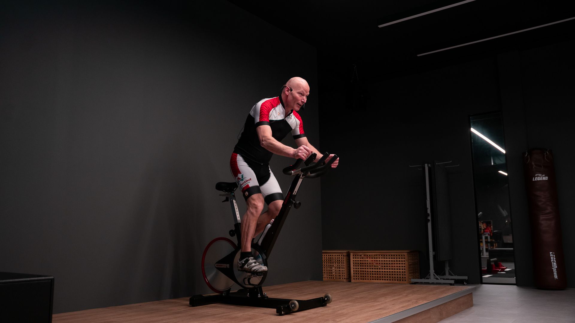 Een man in fietskleding zit op een hometrainer, binnen. Hij is aan het sporten in een donkere sportschool.