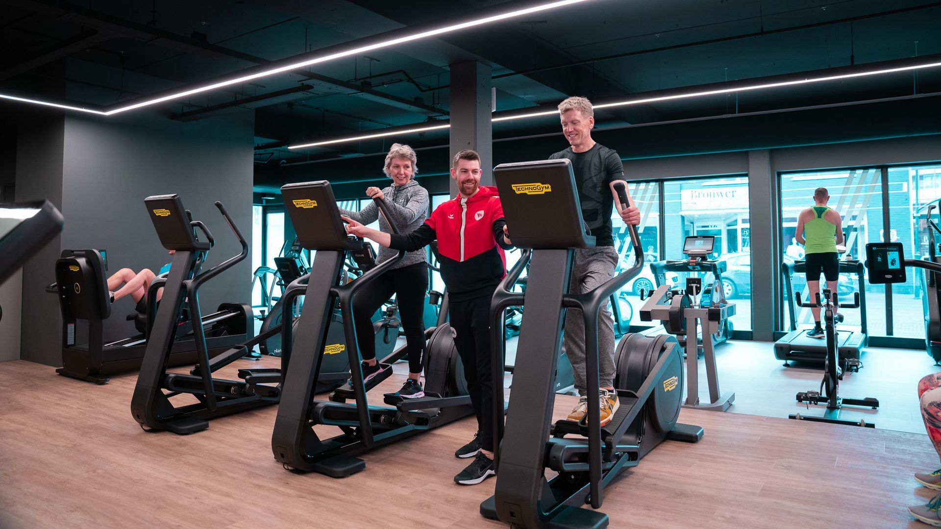 Drie mensen in de sportschool: twee op de crosstrainer, een trainer tussen hen in, anderen op de loopband op de achtergrond.
