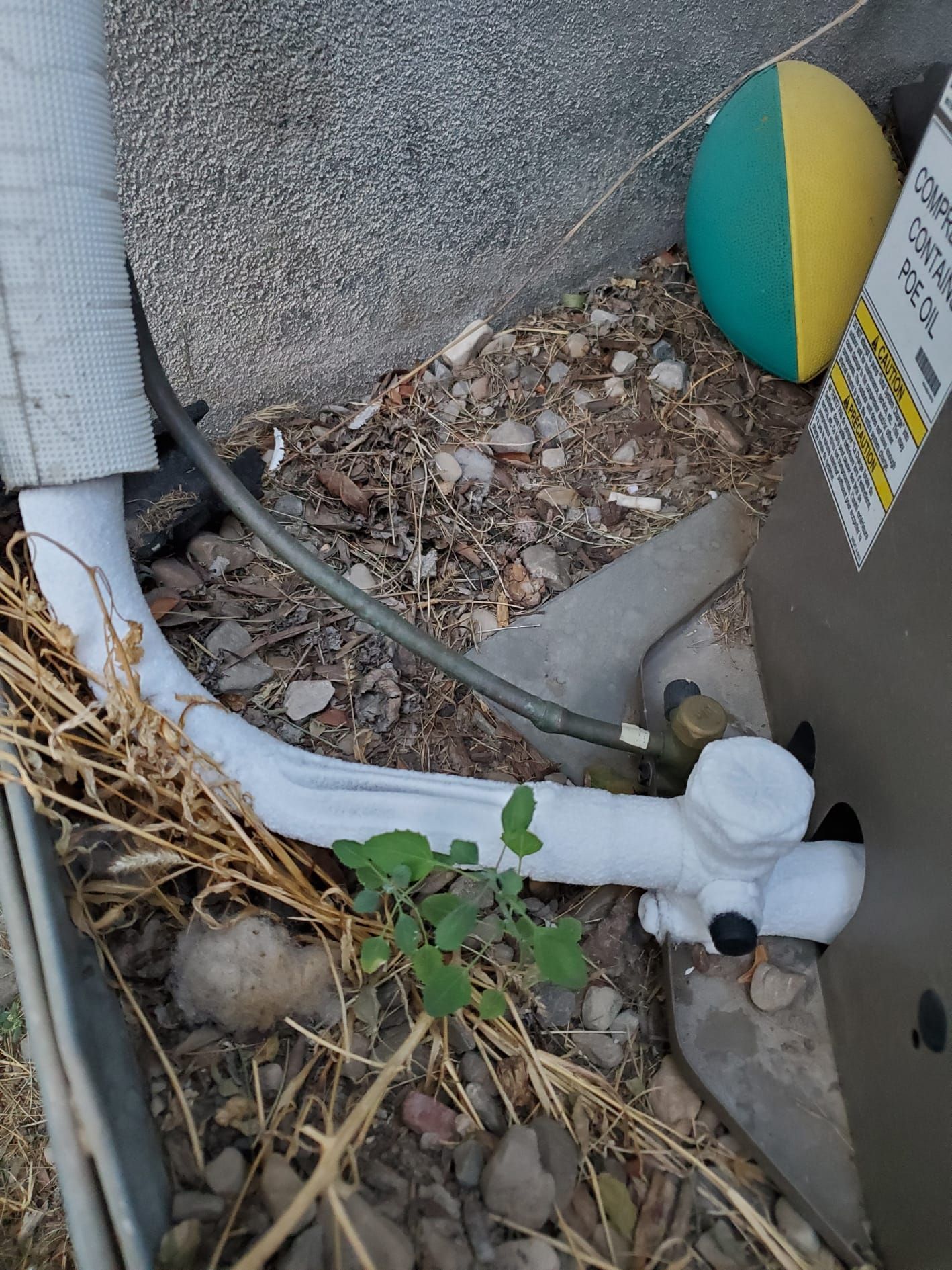 AC unit with insulated pipes, weeds, and a partially visible ball.