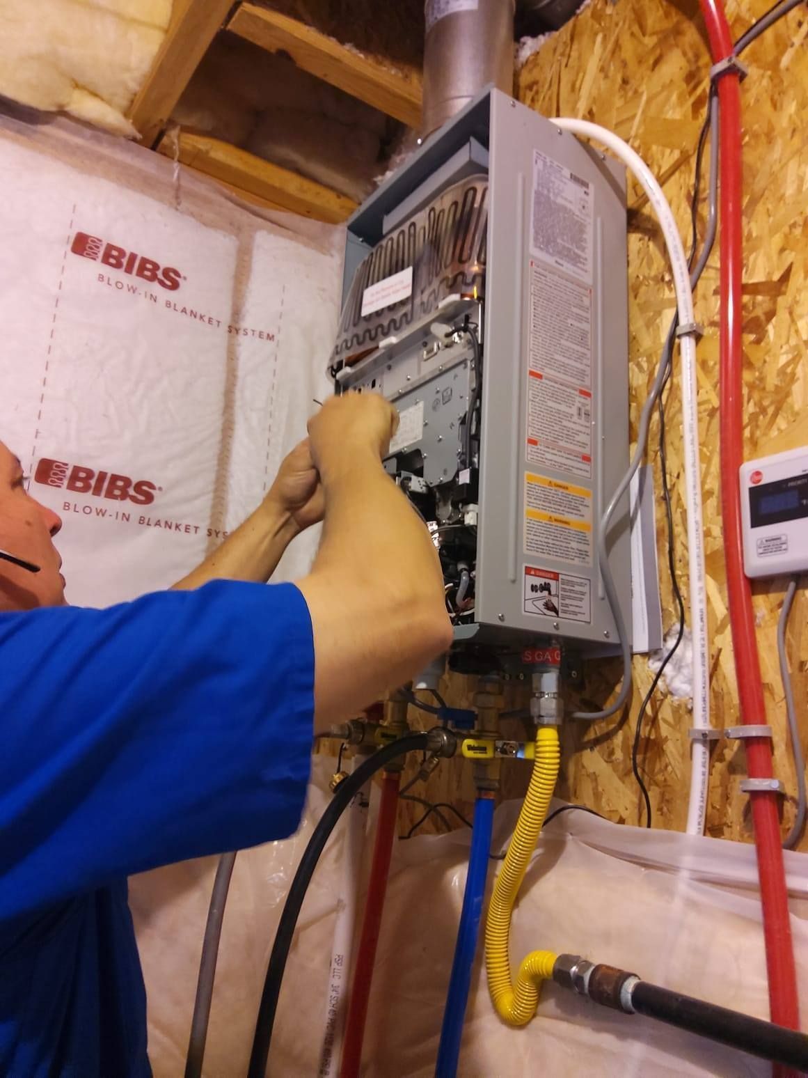 Man working on a tankless water heater, inside a utility room. Yellow gas line and red and white pipes visible.