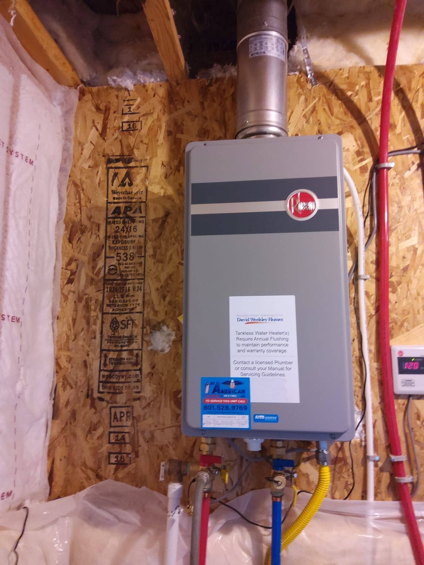 Rheem tankless water heater, mounted on wood paneling, with exhaust pipe, water lines, and safety label.
