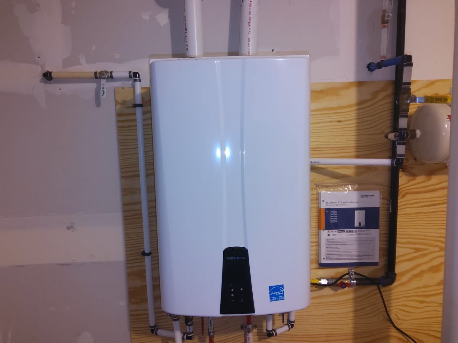 White, wall-mounted tankless water heater with pipes against a wood-paneled wall.