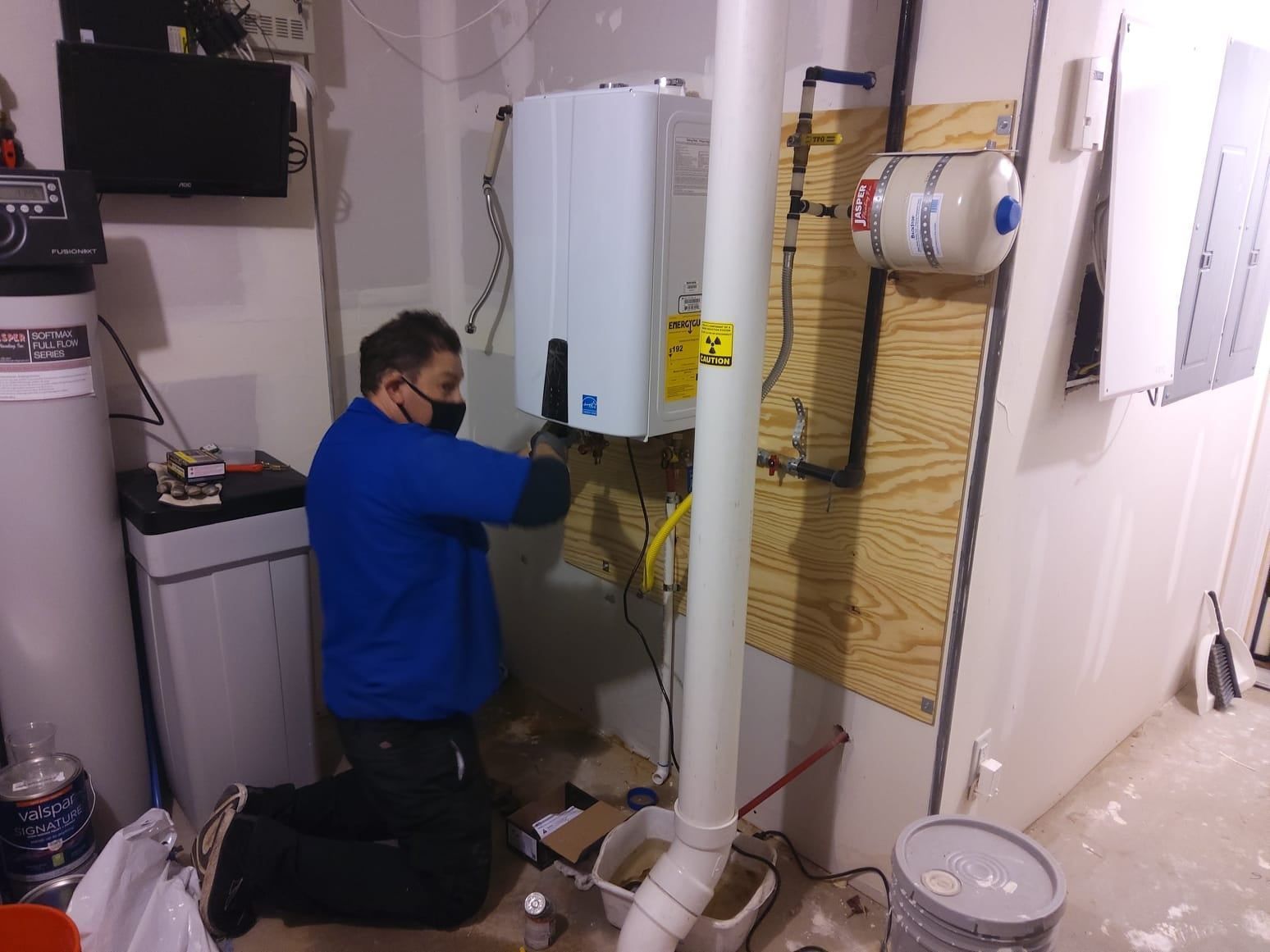 Person installing a tankless water heater in a basement. The person is kneeling, wearing a mask.