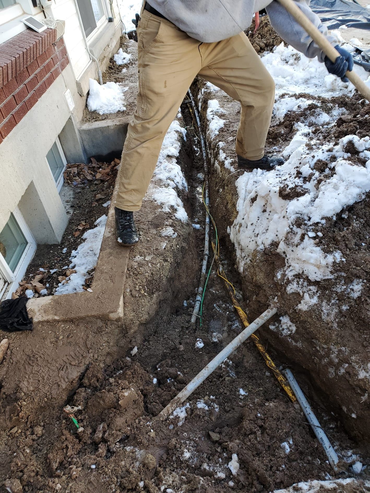 Person digging a trench near a building with some snow on the ground; several pipes visible.