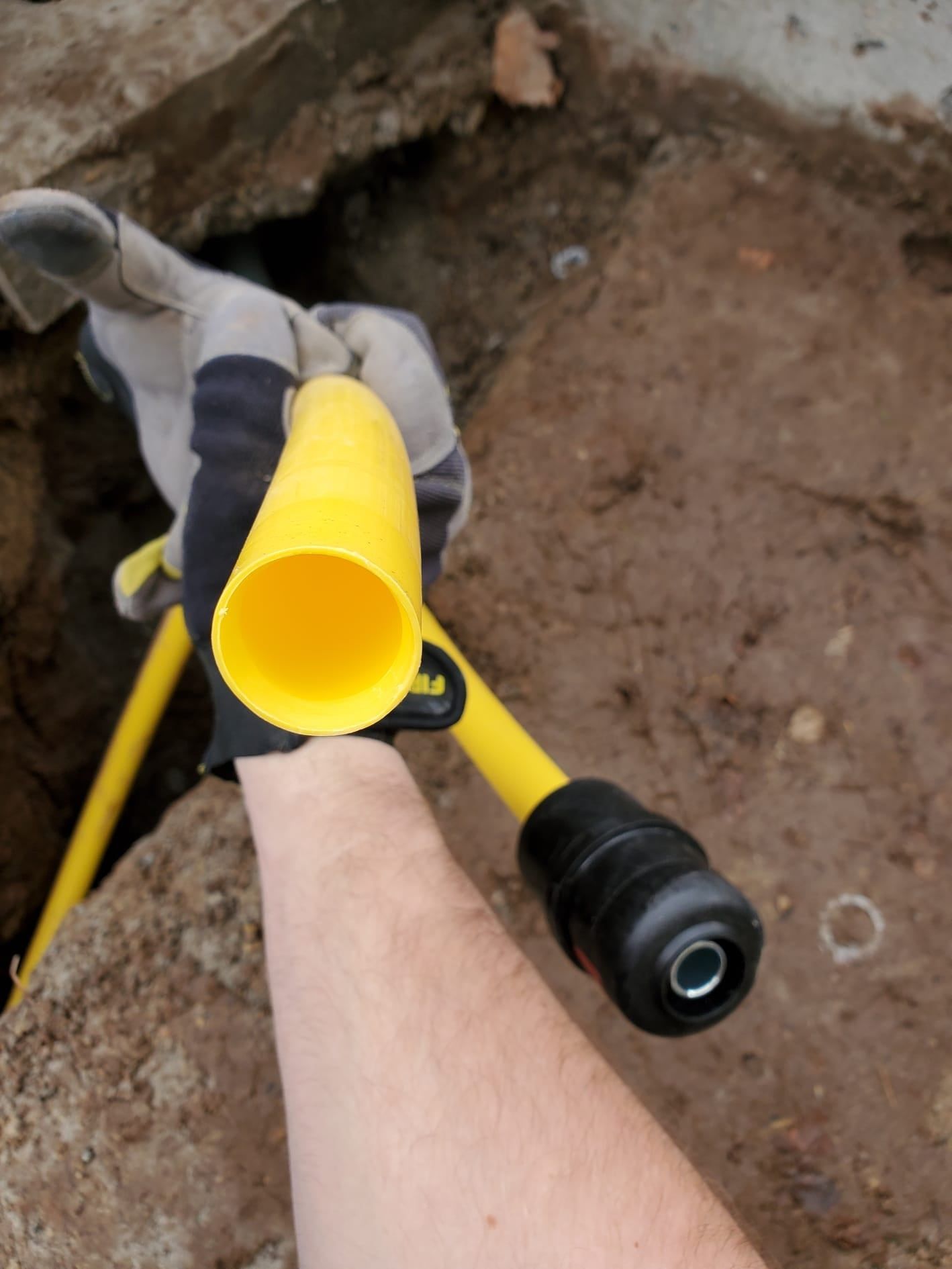 Hand holding a yellow pipe in an excavated area. A gloved hand and a portion of arm are visible.