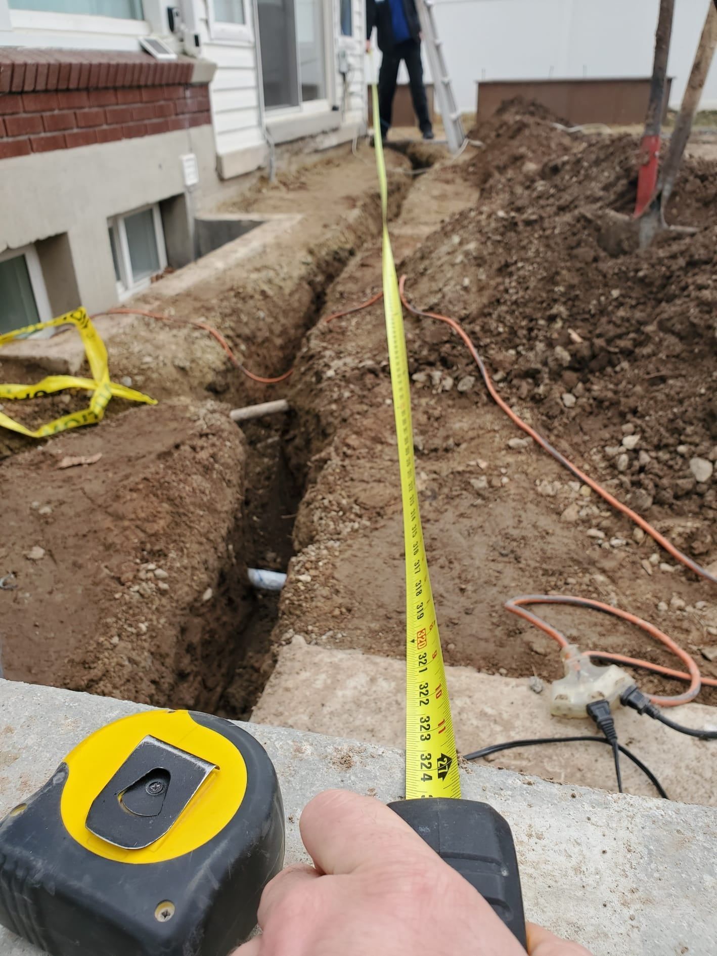 Person measuring a trench with a tape measure; construction site near a building.