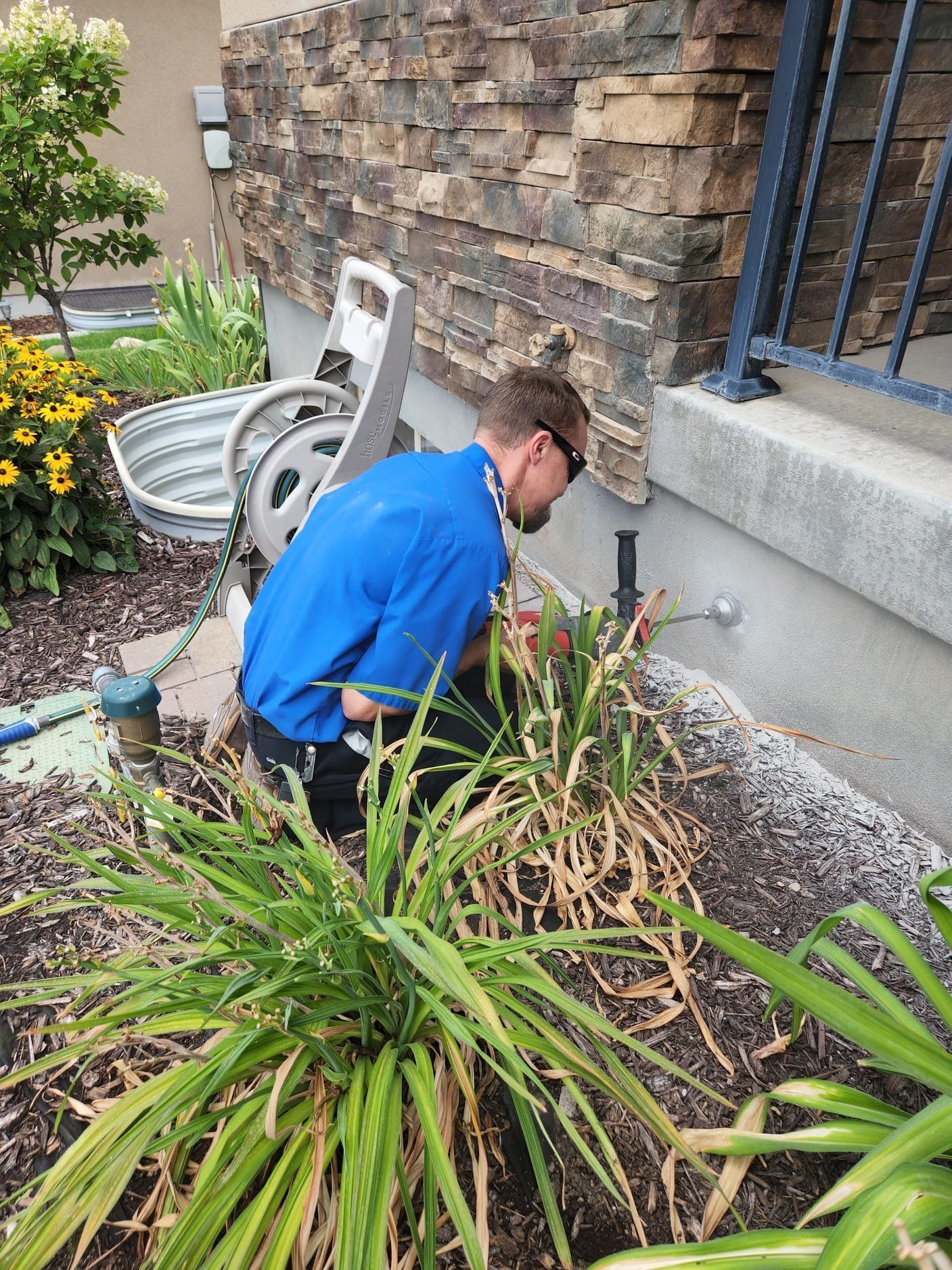 Man in blue shirt, kneeling, working on sprinkler system near house with stone facade and plants.