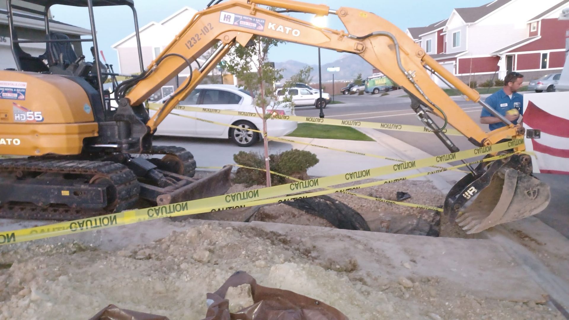 Yellow excavator digging in a residential area, surrounded by caution tape. A worker stands nearby.