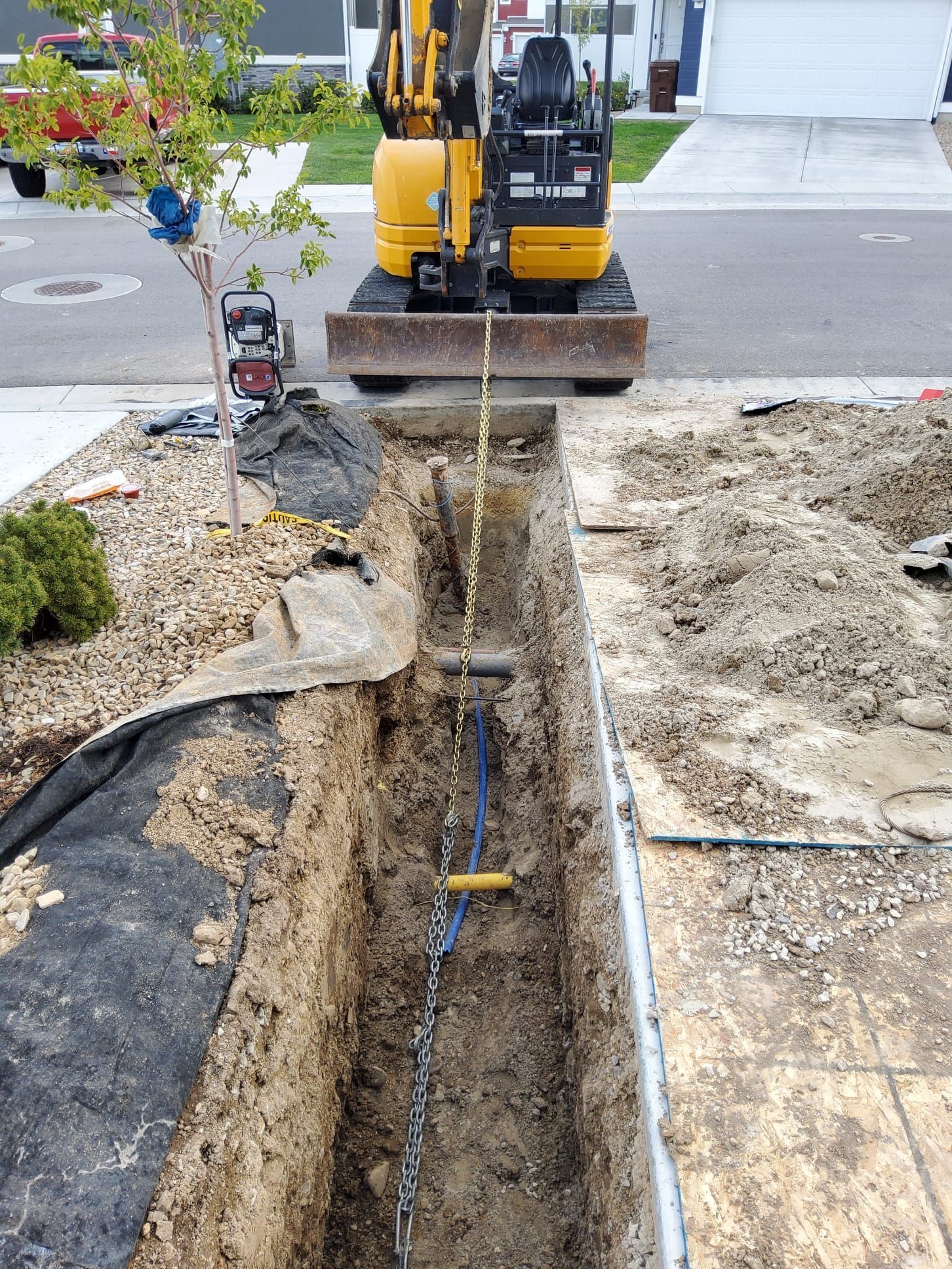Mini excavator digging trench in a residential area, chain and buried object visible.