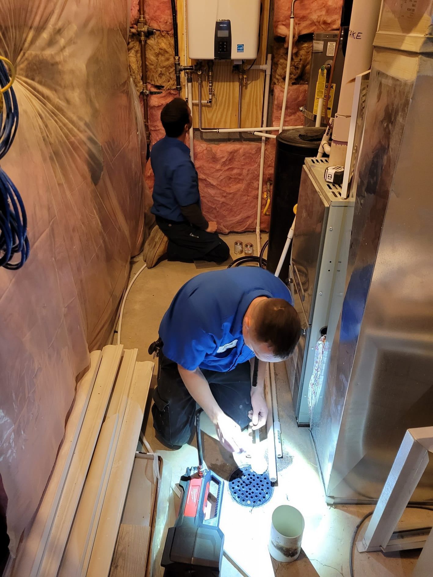 Two men in blue shirts are working on plumbing in a basement, one kneeling, using tools, the other looking up.