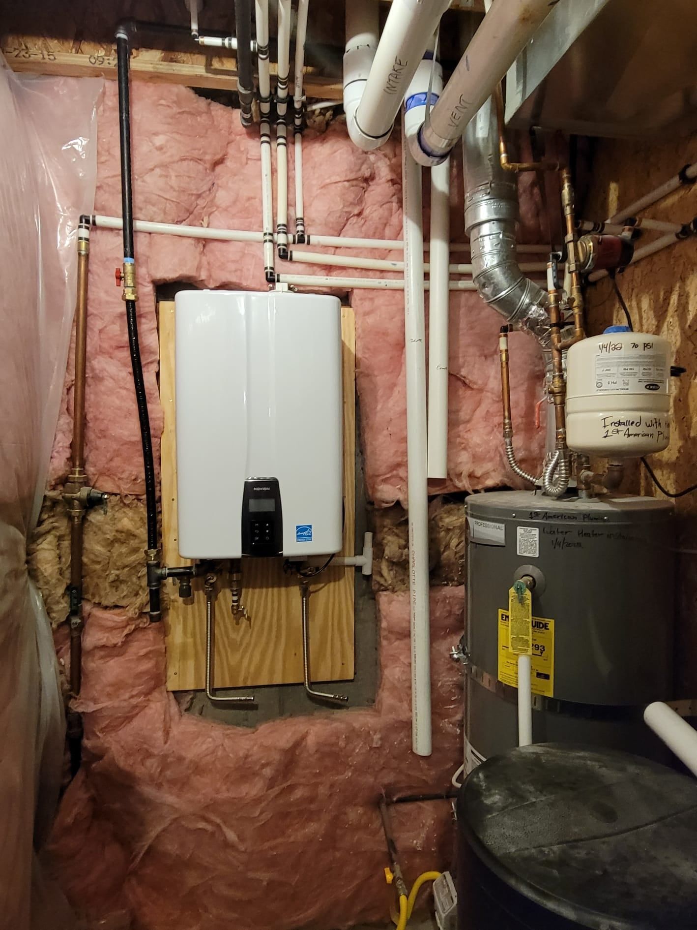 Water heaters and plumbing in a basement, with pink insulation and pipes on the wall.