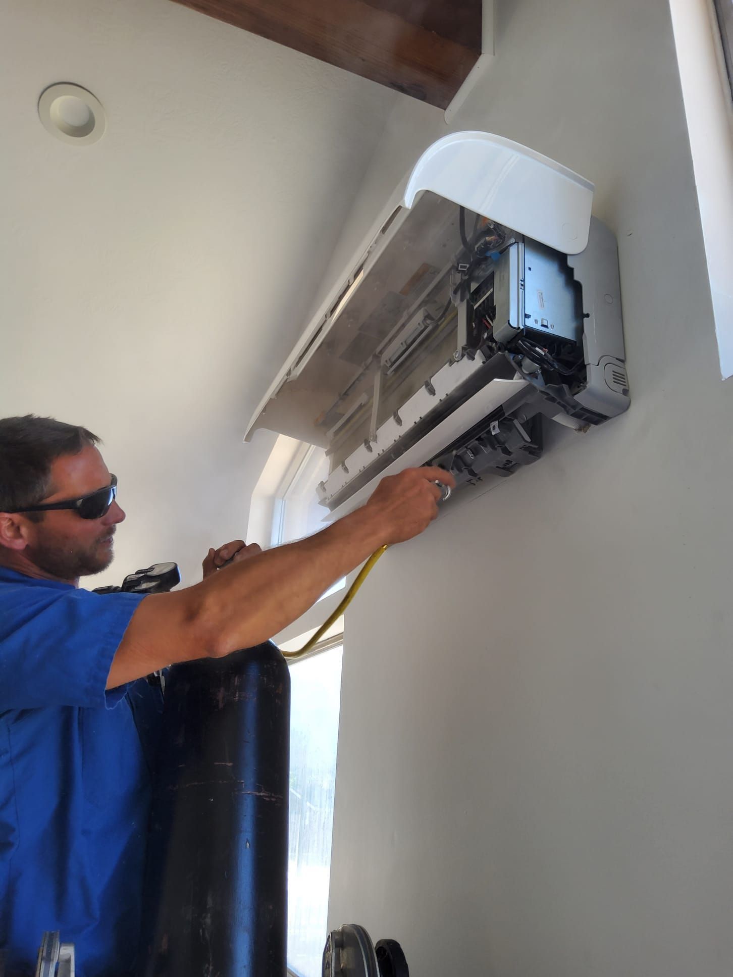 Man in blue shirt services an air conditioning unit on a wall, holding a tool near it.
