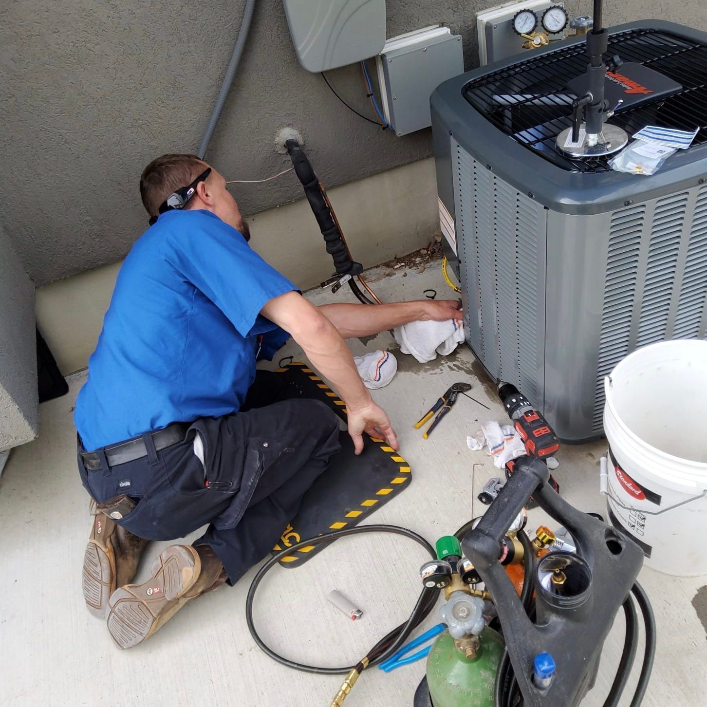 HVAC technician working on an air conditioning unit outside, wearing a blue shirt and safety glasses.