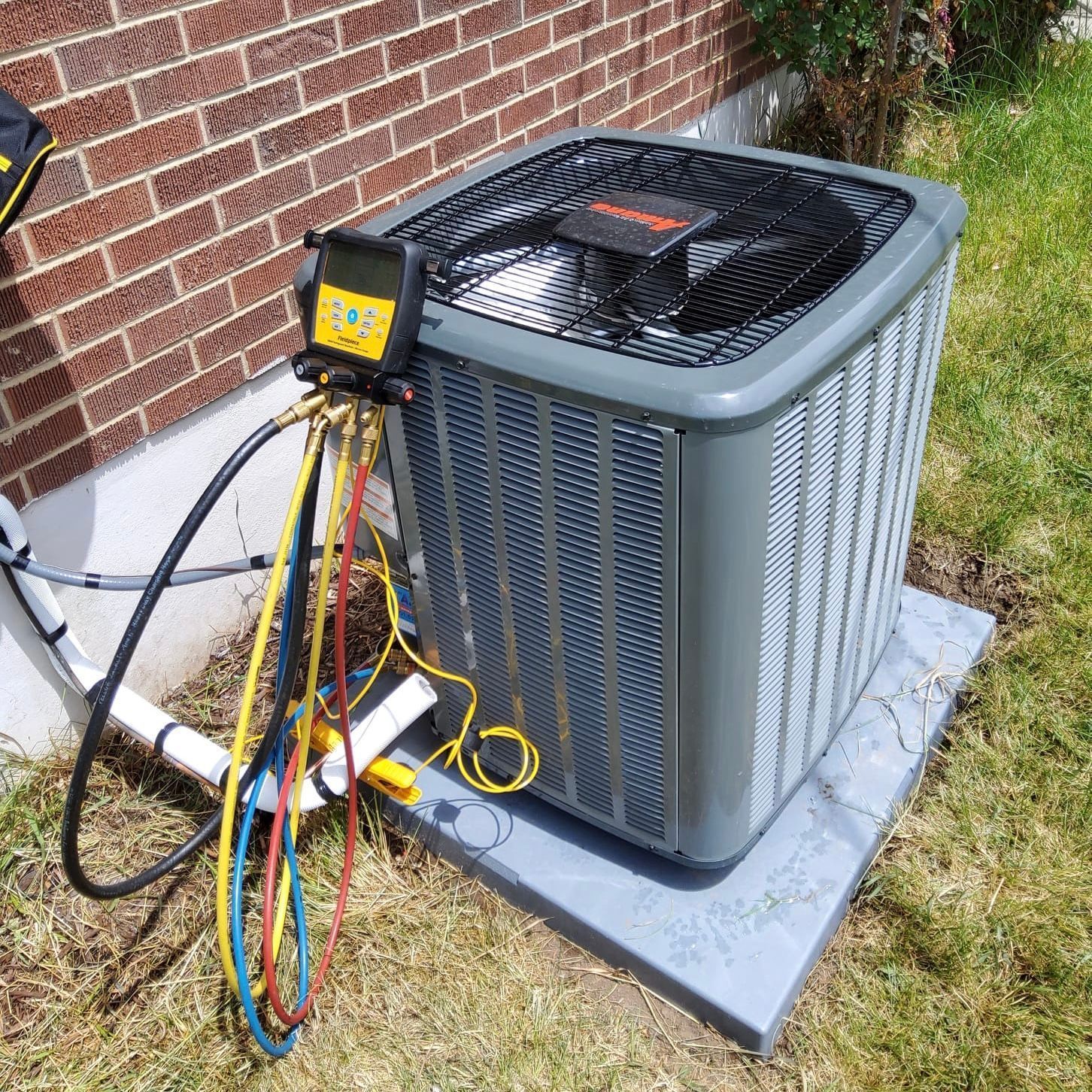 HVAC unit with gauges and hoses connected. Gray outdoor air conditioner on concrete pad, near brick wall and grass.