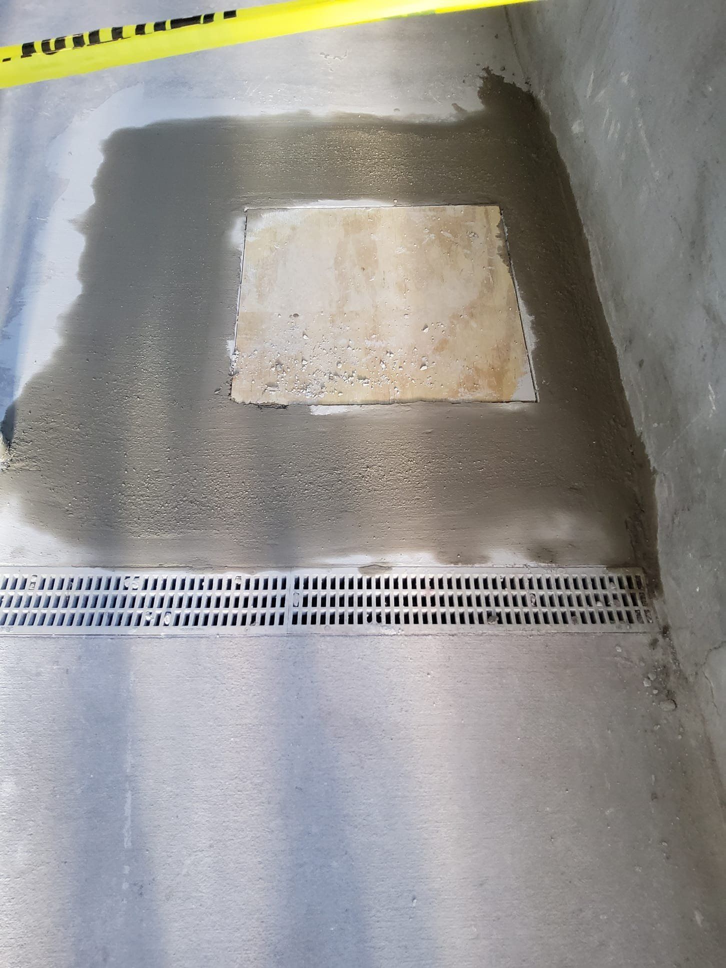 Concrete floor with a rectangular metal drain and a patch of wet concrete near a yellow caution tape.