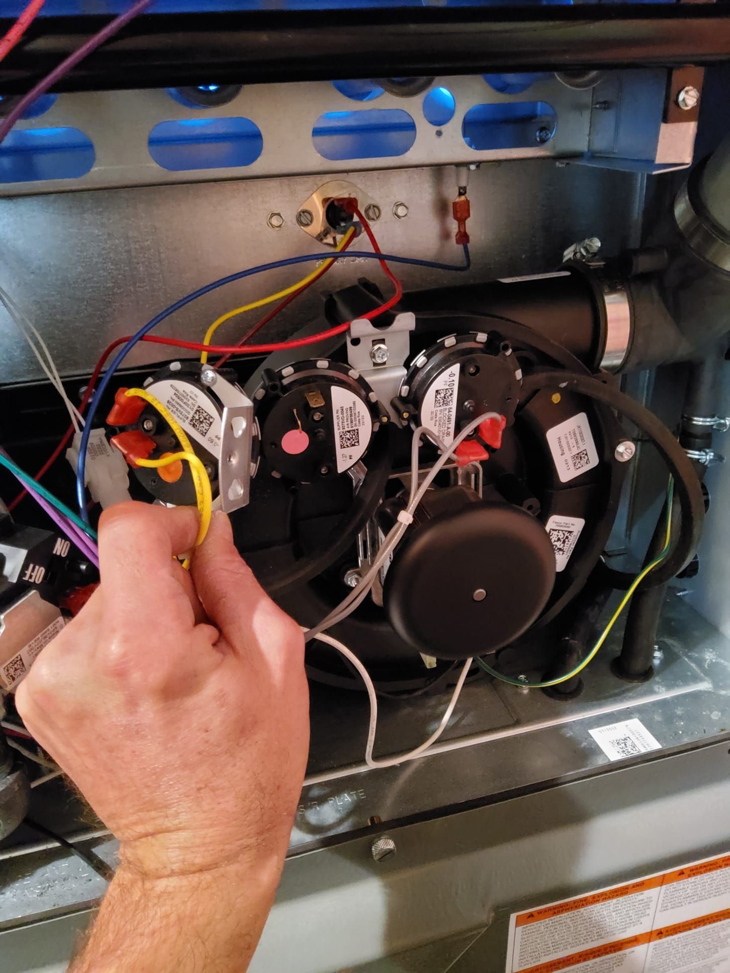 Hand holding a yellow wire in a furnace. The furnace interior has wires, components, and a black blower.