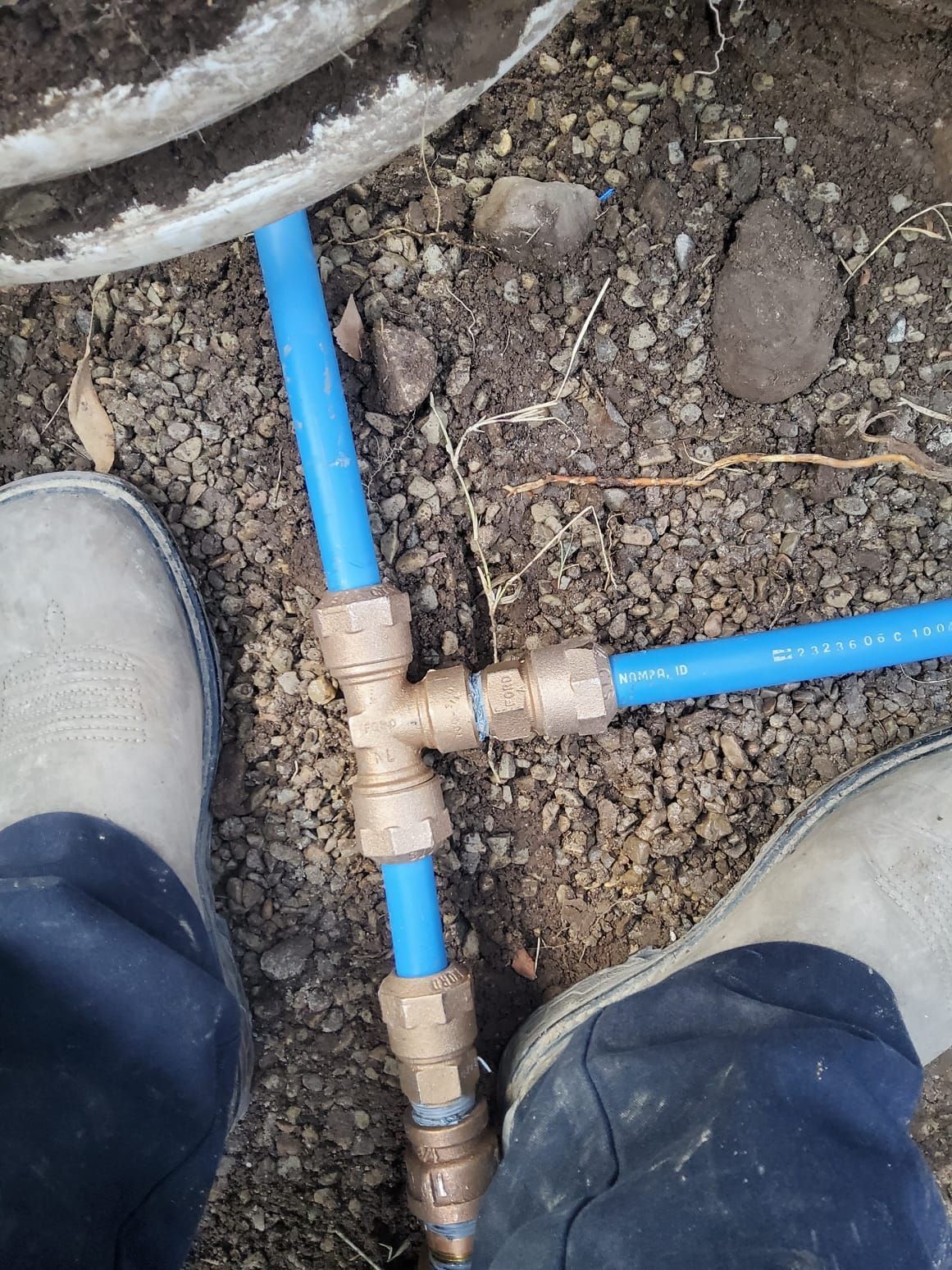 Blue PEX pipes connected with brass fittings buried in soil. A person's legs and boots are in the frame.