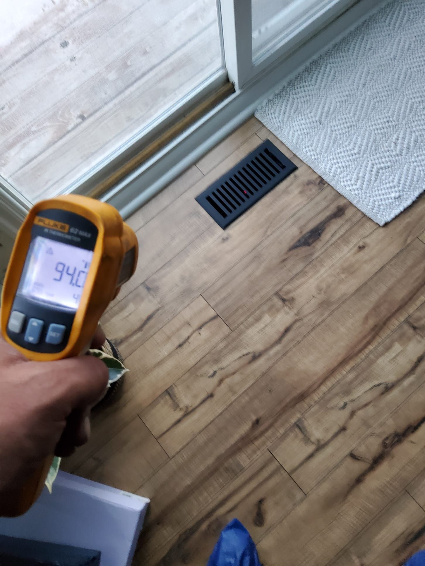 A hand holds a yellow thermometer pointed at a brown floor, reading 94 degrees. A vent and sliding door are in the background.