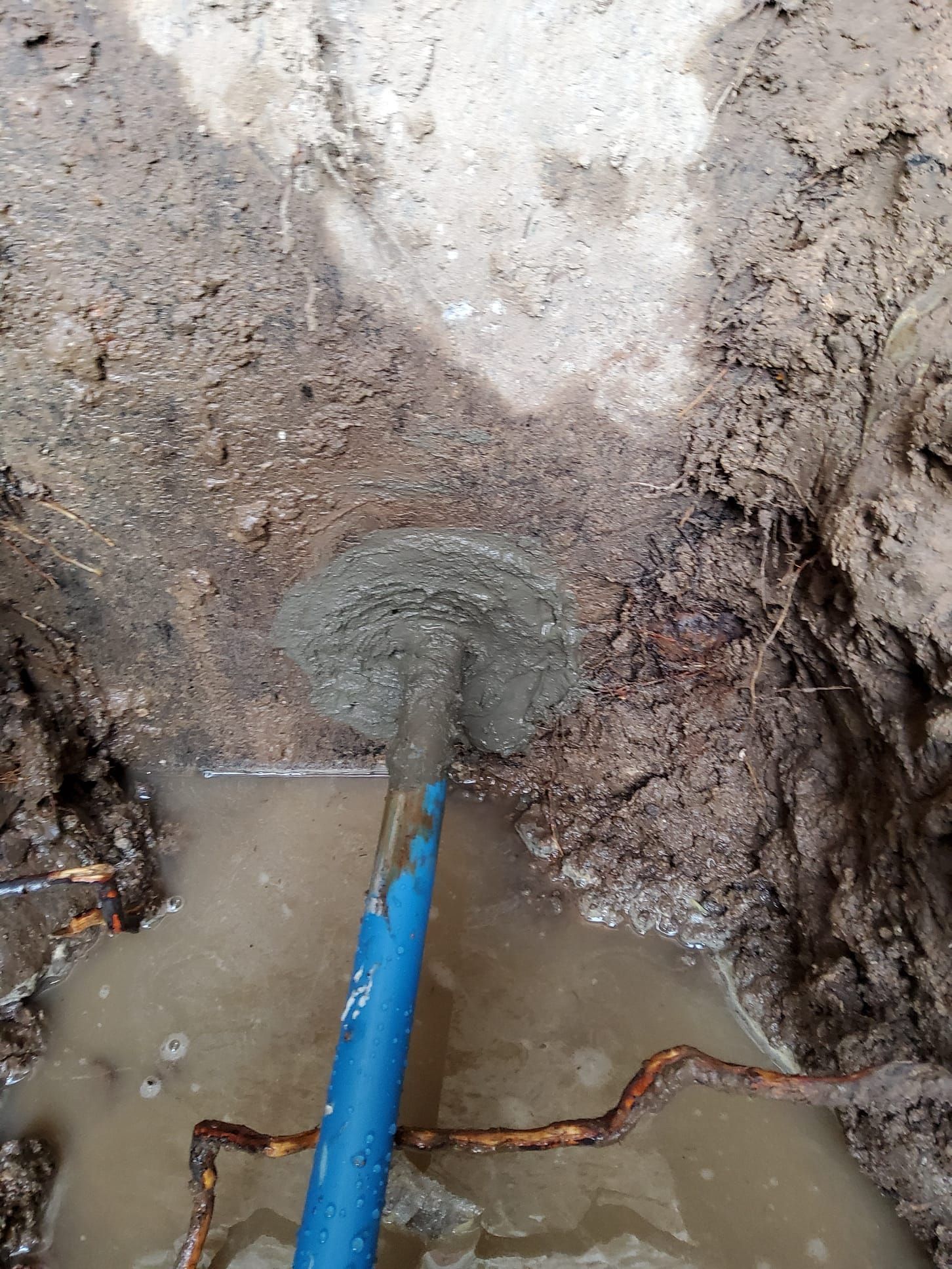 Shovel in a hole filled with mud and water; blue handle, brown soil, and a rusty pipe are visible.