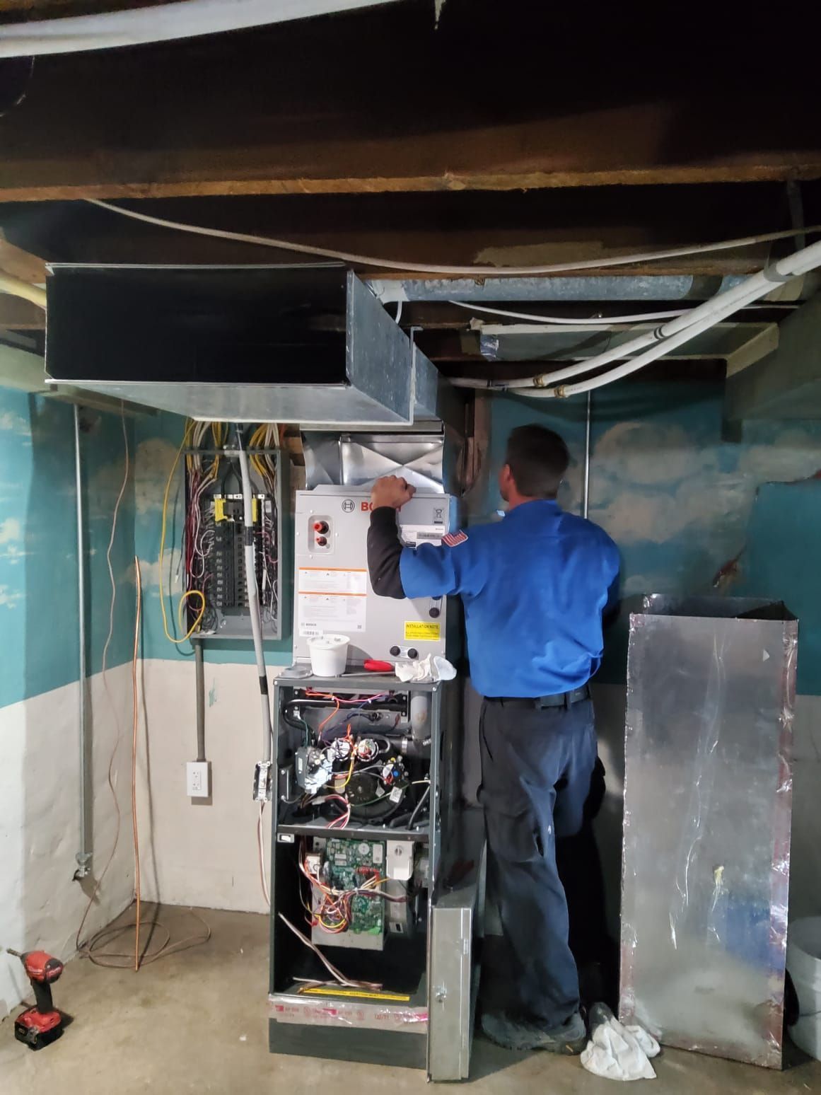 HVAC technician inspecting a furnace in a basement. The technician is wearing a blue shirt.