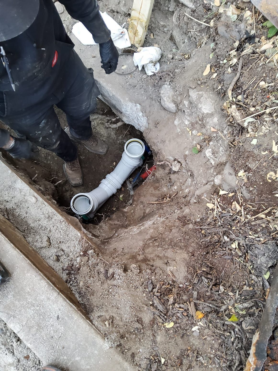 Person installing a gray drain pipe in a trench, next to a concrete structure.