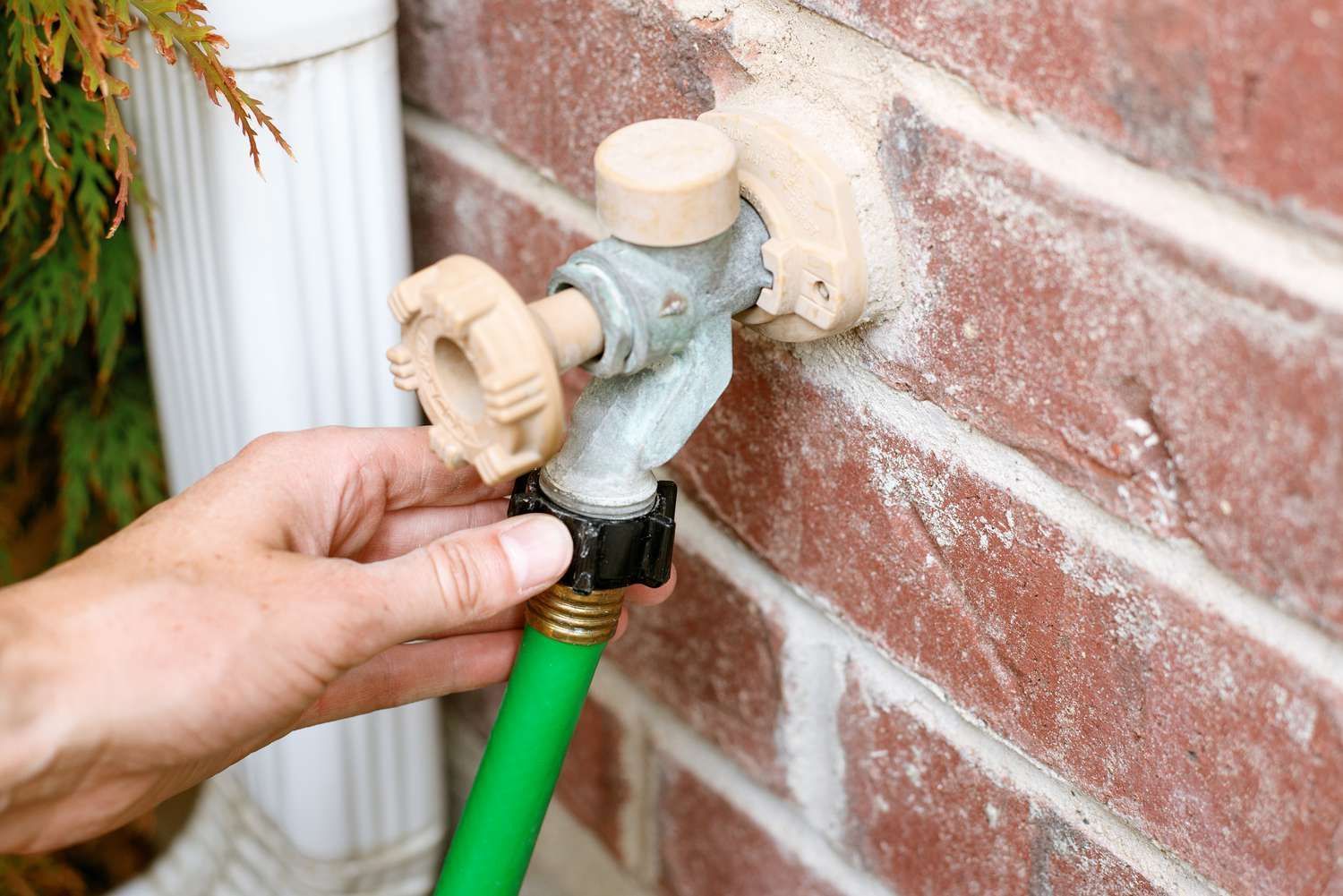 Hand connecting a green hose to an outdoor water faucet on a brick wall.