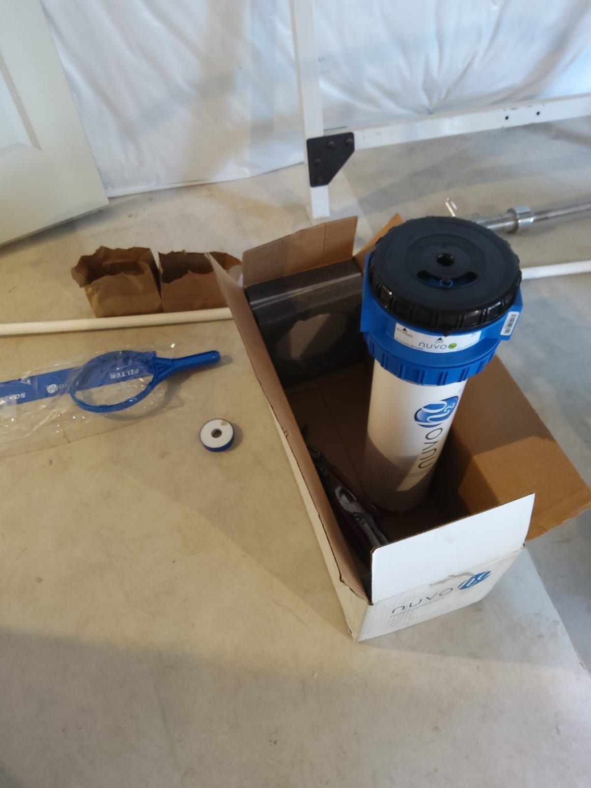 Water filter components in an open cardboard box on a concrete floor near a white wall and plumbing pipes.