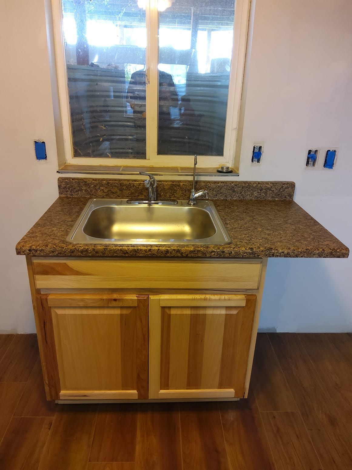 Small sink and cabinet installed beneath a window. Brown countertop, stainless steel sink, and light wood cabinet.