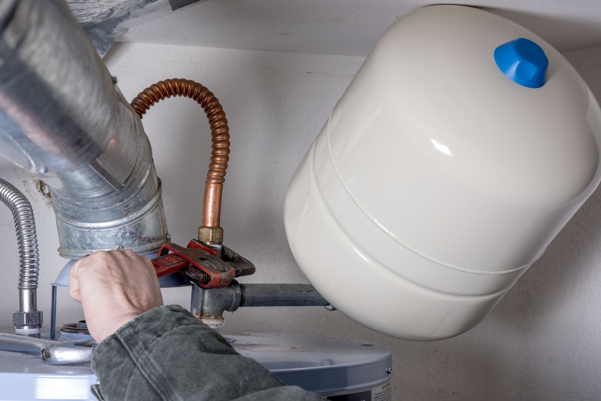 Person using a wrench to connect a white expansion tank to a water heater in a utility room.