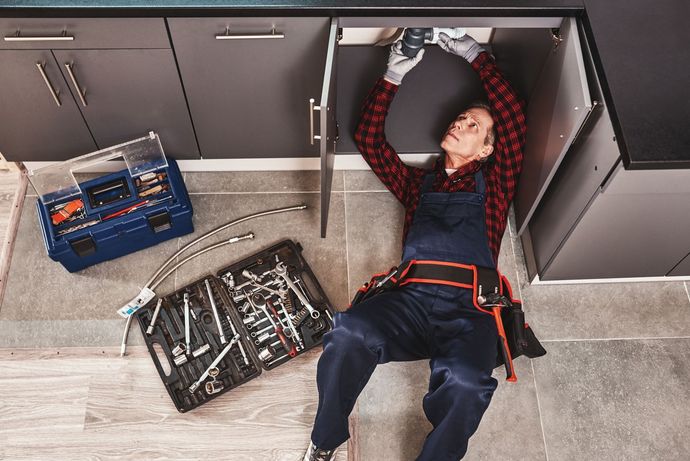 Person lying on a kitchen floor beside open toolboxes and cabinets, reaching overhead while doing repairs