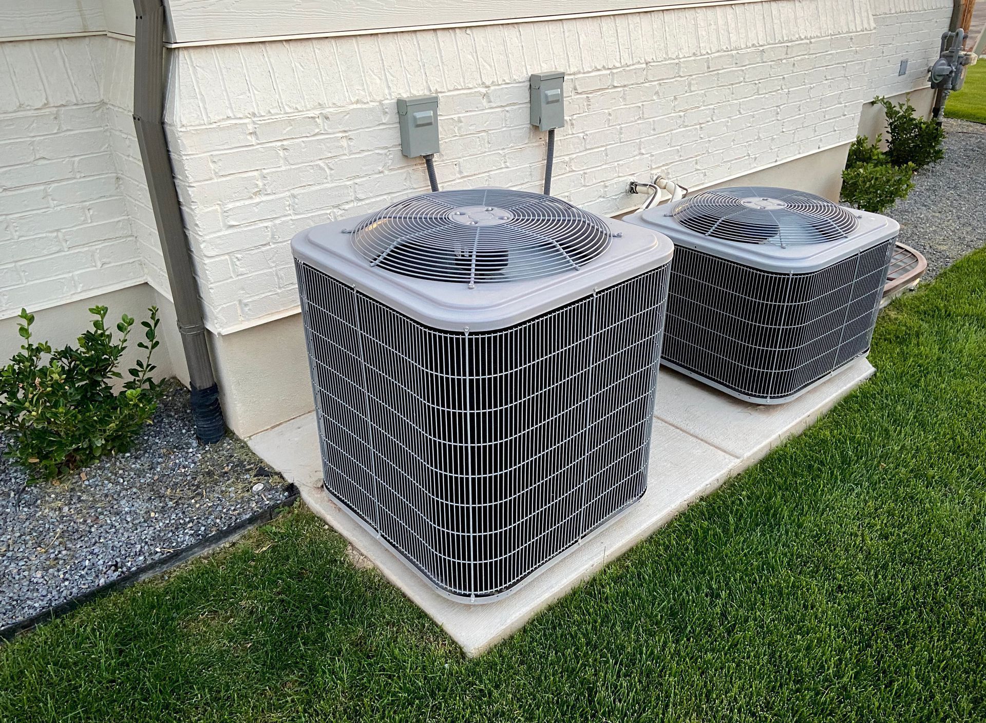 Two air conditioning units on a concrete pad next to a building, with electrical boxes above them, and grass around them.