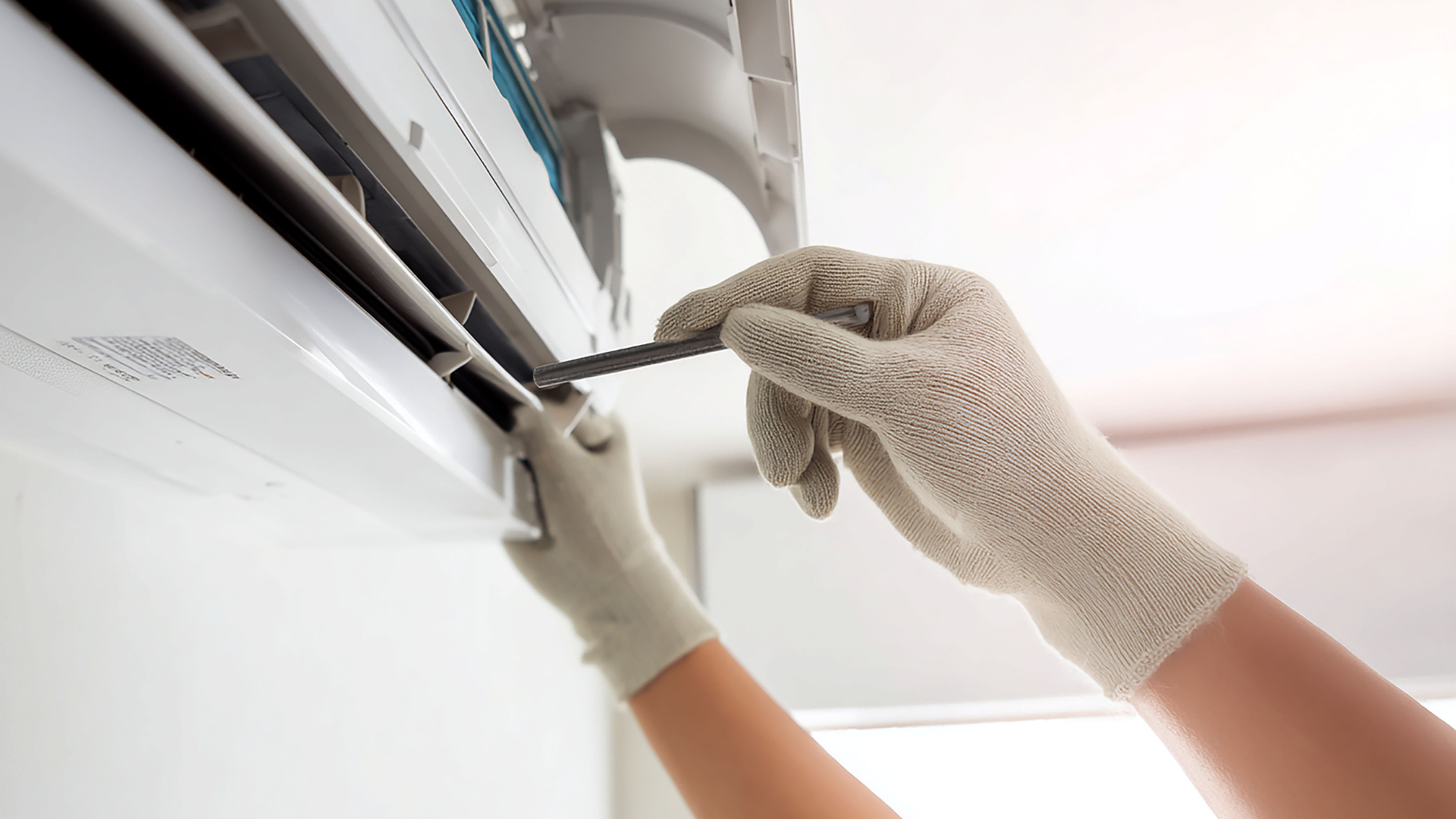 Person in gloves using a tool to repair an air conditioner mounted on a white wall.