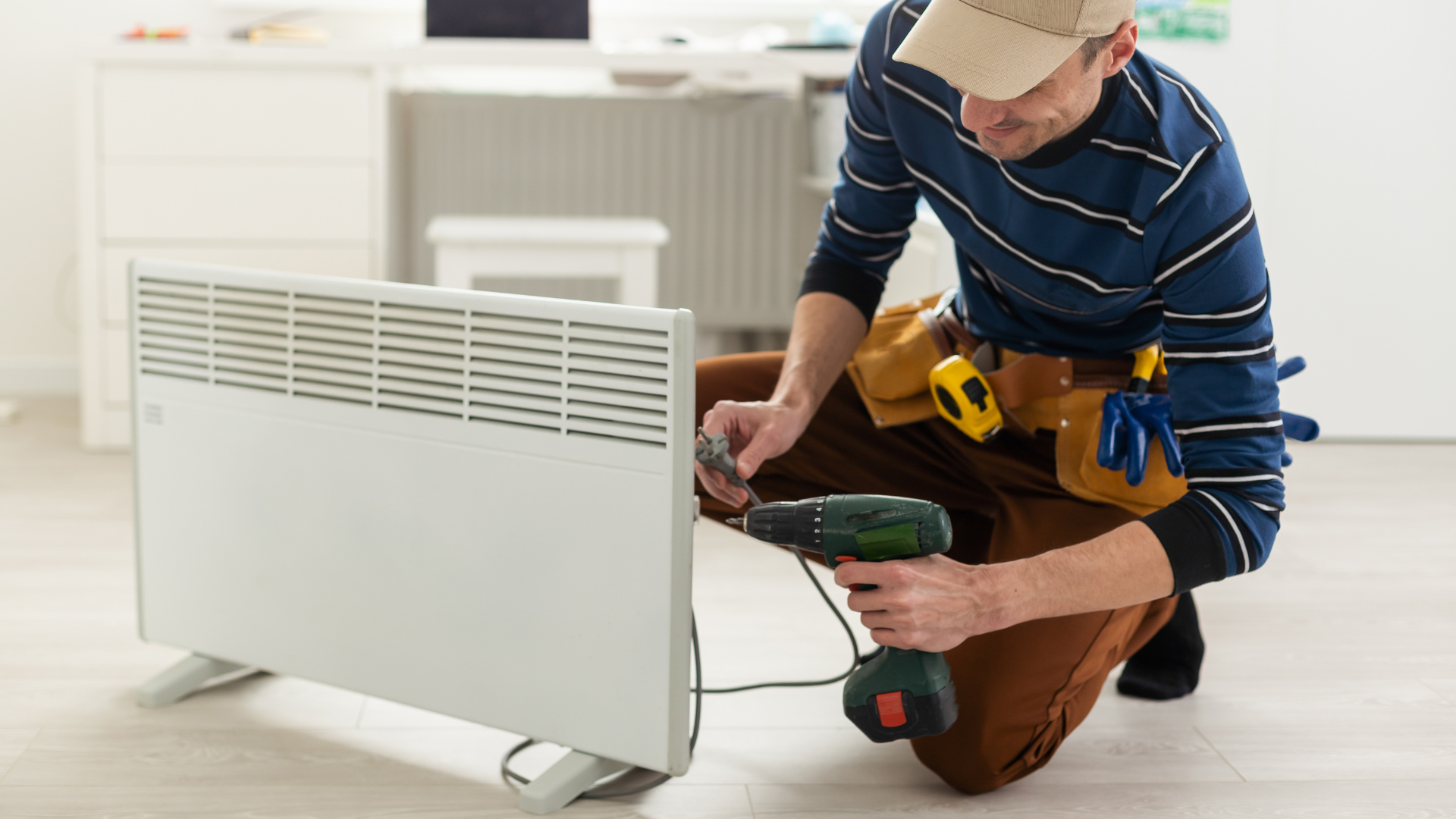 Man in work clothes uses a power drill on a white space heater in a room.