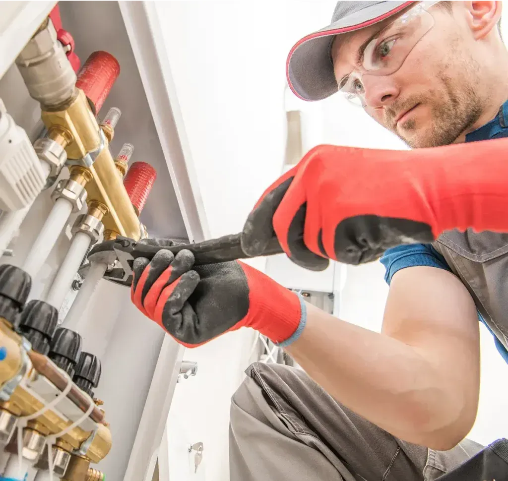 Plumber in safety glasses and gloves using a wrench on pipes within a utility panel.