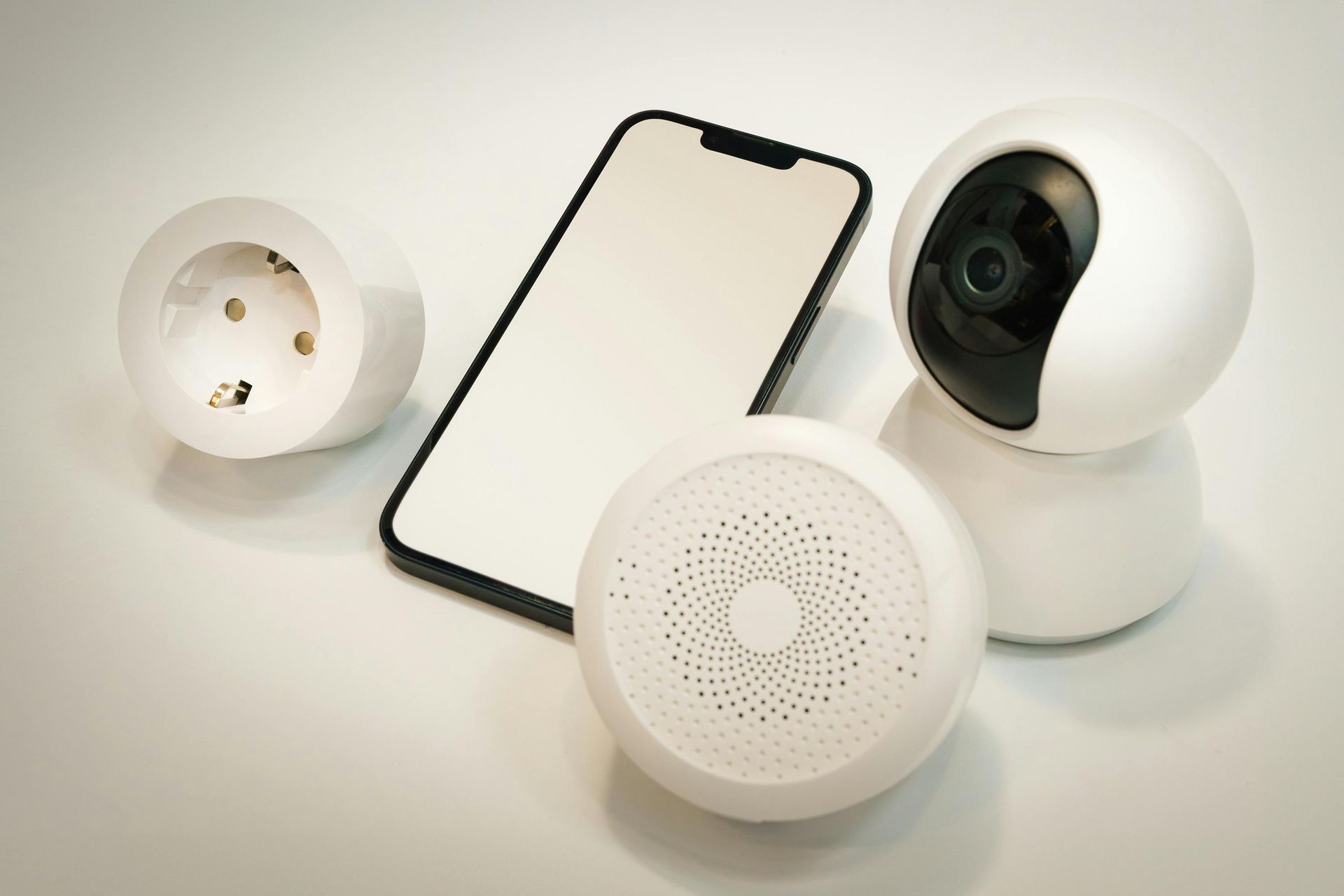 A smartphone, security camera, smart speaker, and smart plug on a white surface.