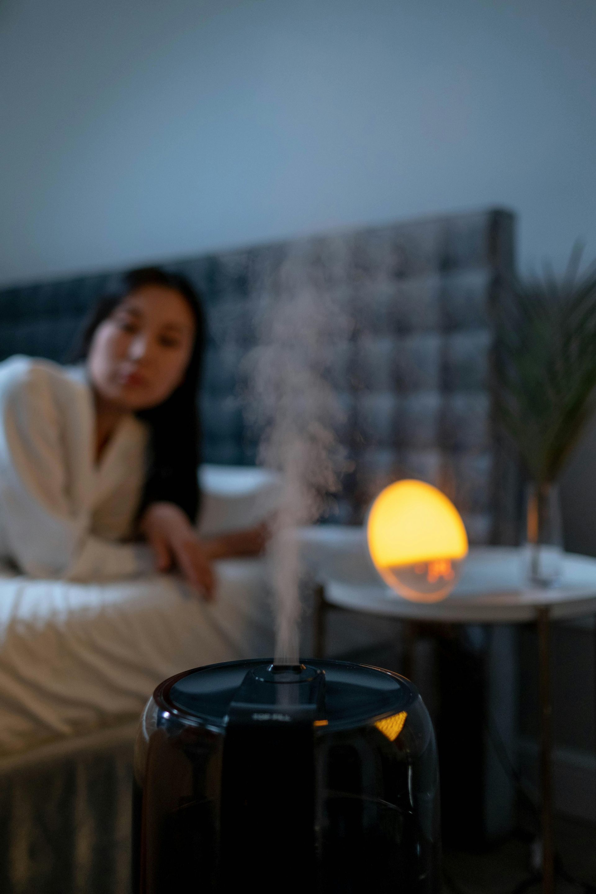 Black humidifier with vapor plume, woman in bed with lit orb on side table.