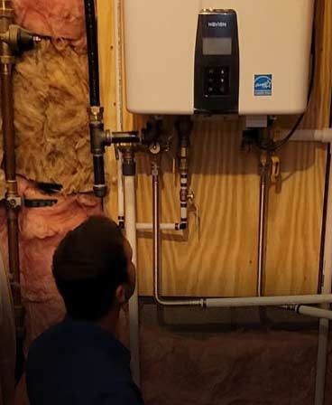 Man inspecting a tankless water heater installed on a wooden wall in a utility room. Copper pipes and insulation visible.
