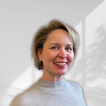 A woman in a grey sweater is smiling in front of a white wall.