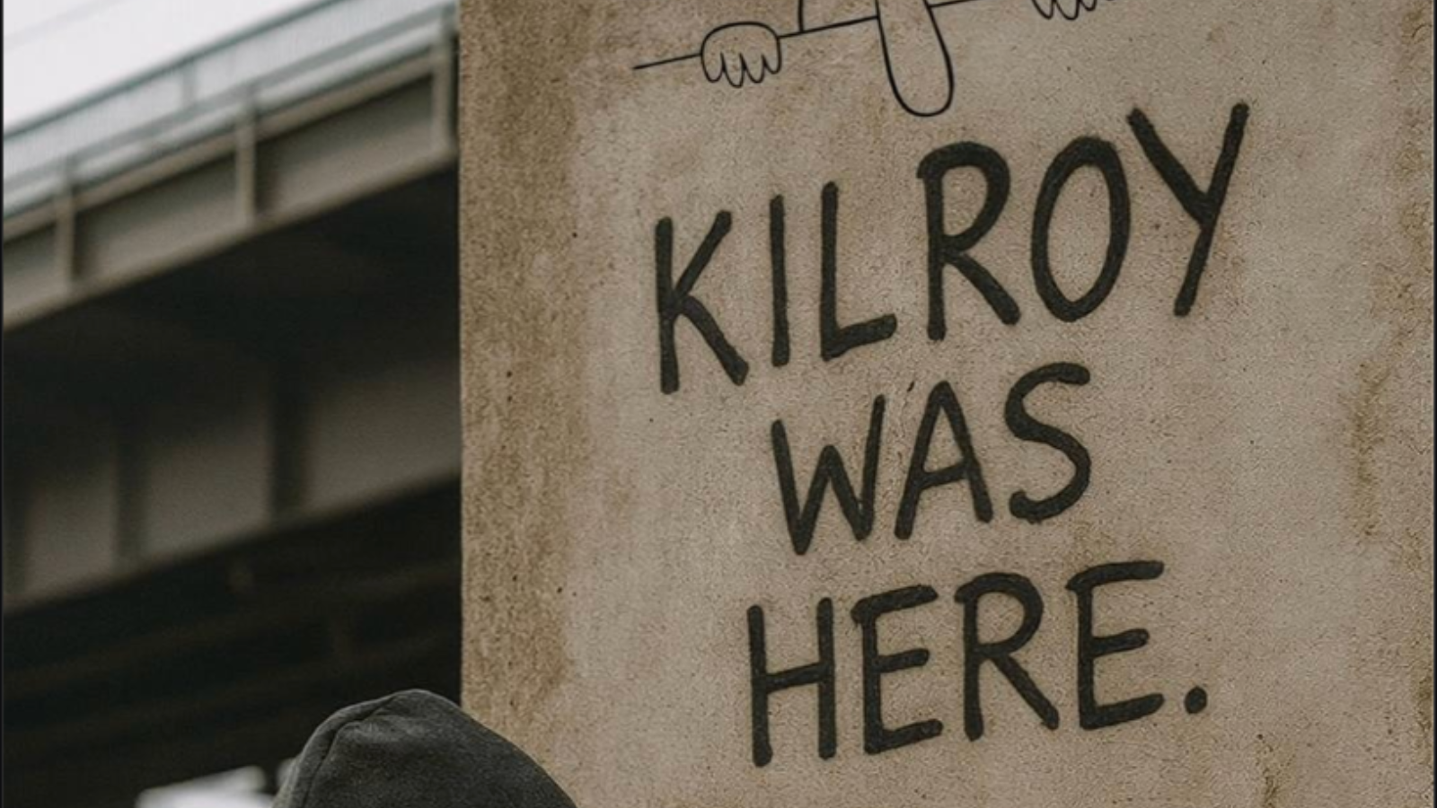 Person spray-painting “Kilroy Was Here” graffiti on a wall beneath the iconic peeking face drawing.