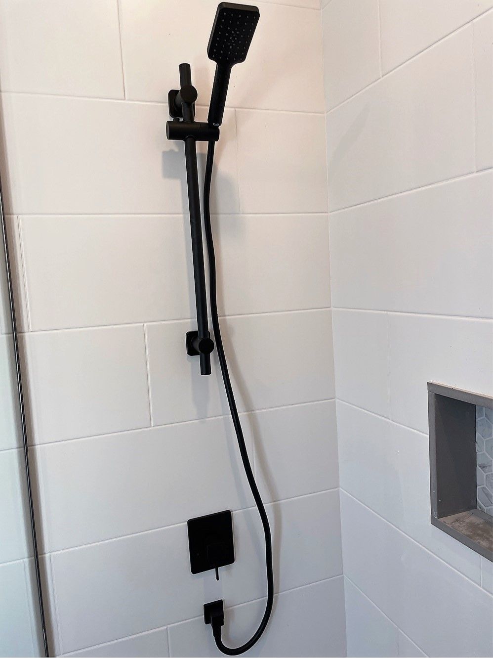 A black shower head is hanging from a white tiled wall in a bathroom.