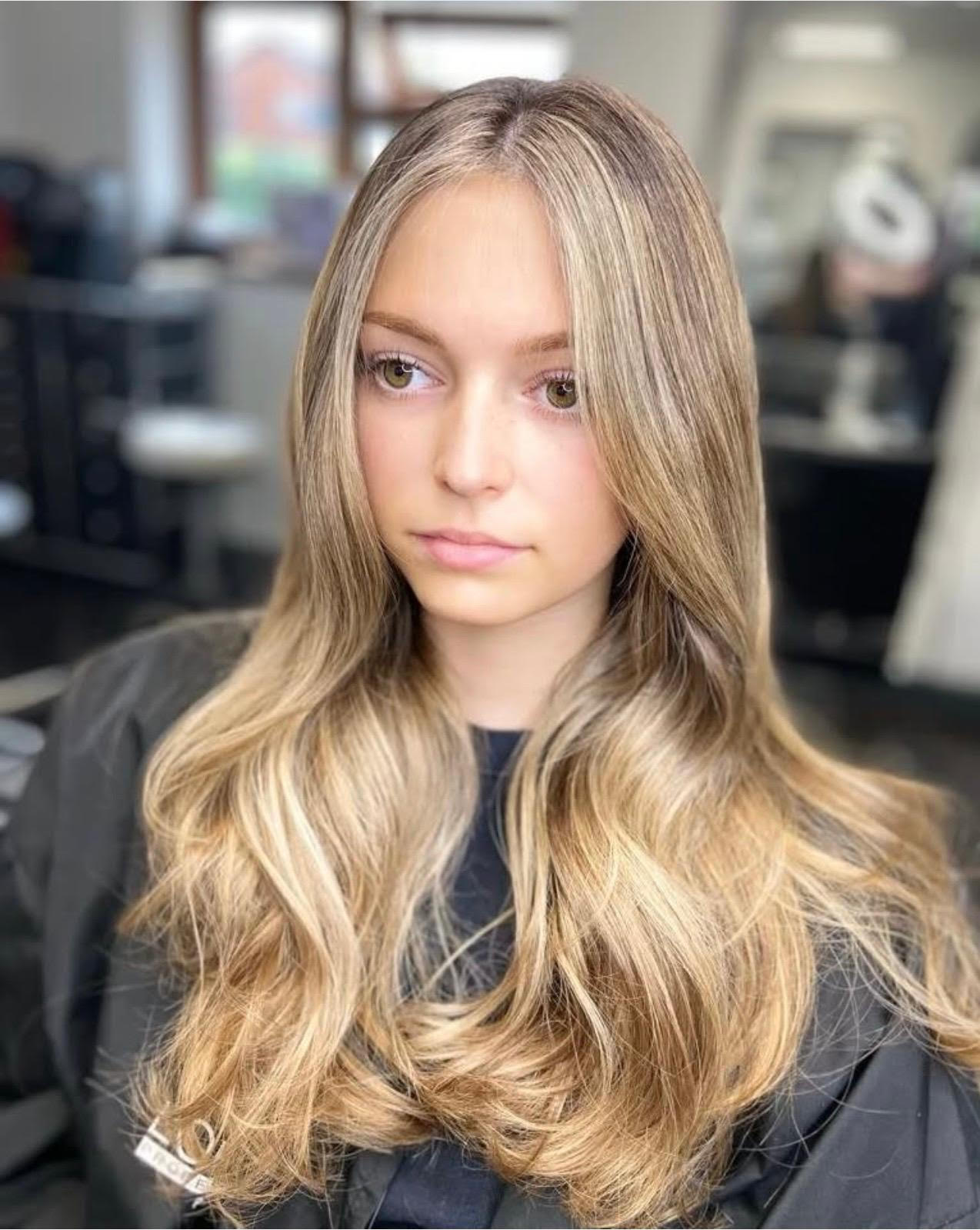 Young person with long blonde hair and soft waves, seated in a salon chair, with a blurred interior background