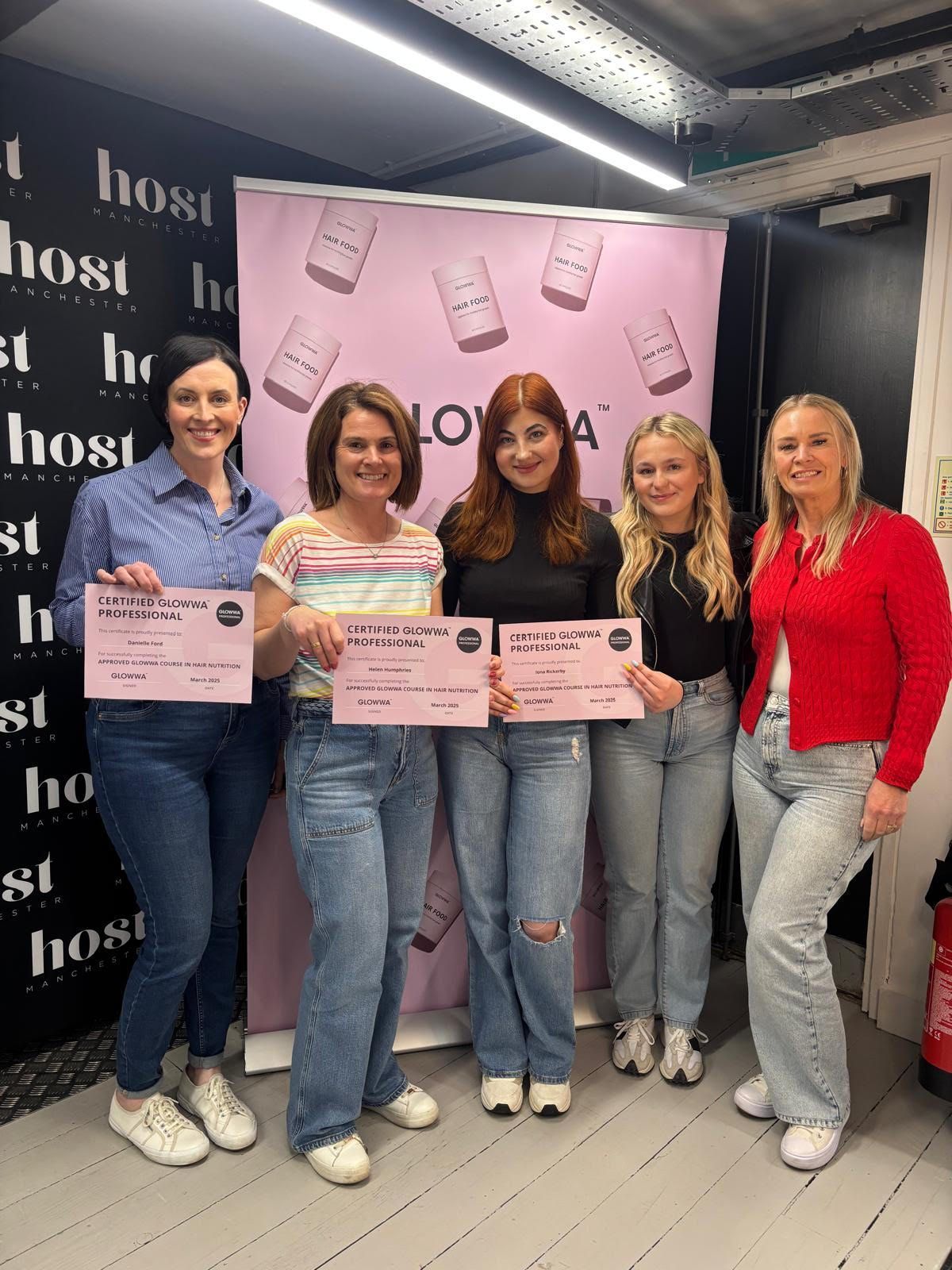 Five women holding certificates pose at a booth with a pink “Hello” backdrop.
