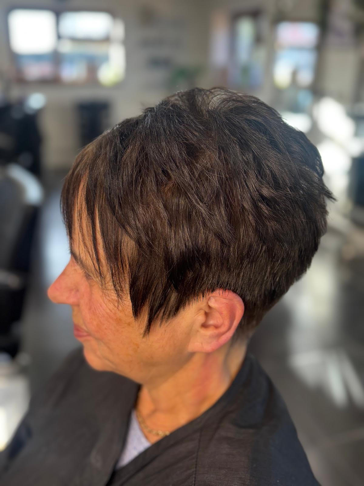 Profile view of a short dark layered haircut in a salon chair, with a blurred salon background