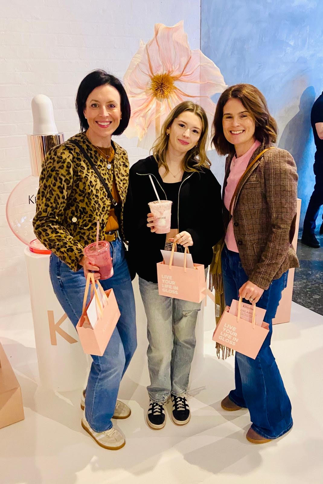 Three people smiling at a boutique event, holding pink shopping bags and drinks.
