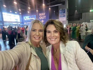 Two women smiling for a selfie at a busy indoor event, with a “WAY OUT” sign in the background.