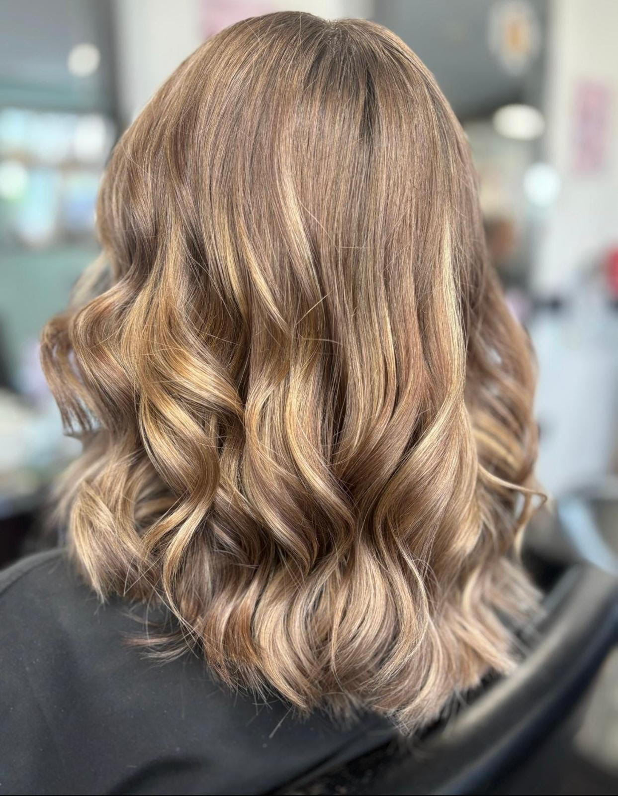 Wavy, shoulder-length brown hair with blonde highlights, styled in loose curls in a salon chair
